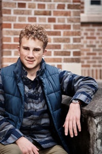 a young man in a blue vest is leaning against a brick wall